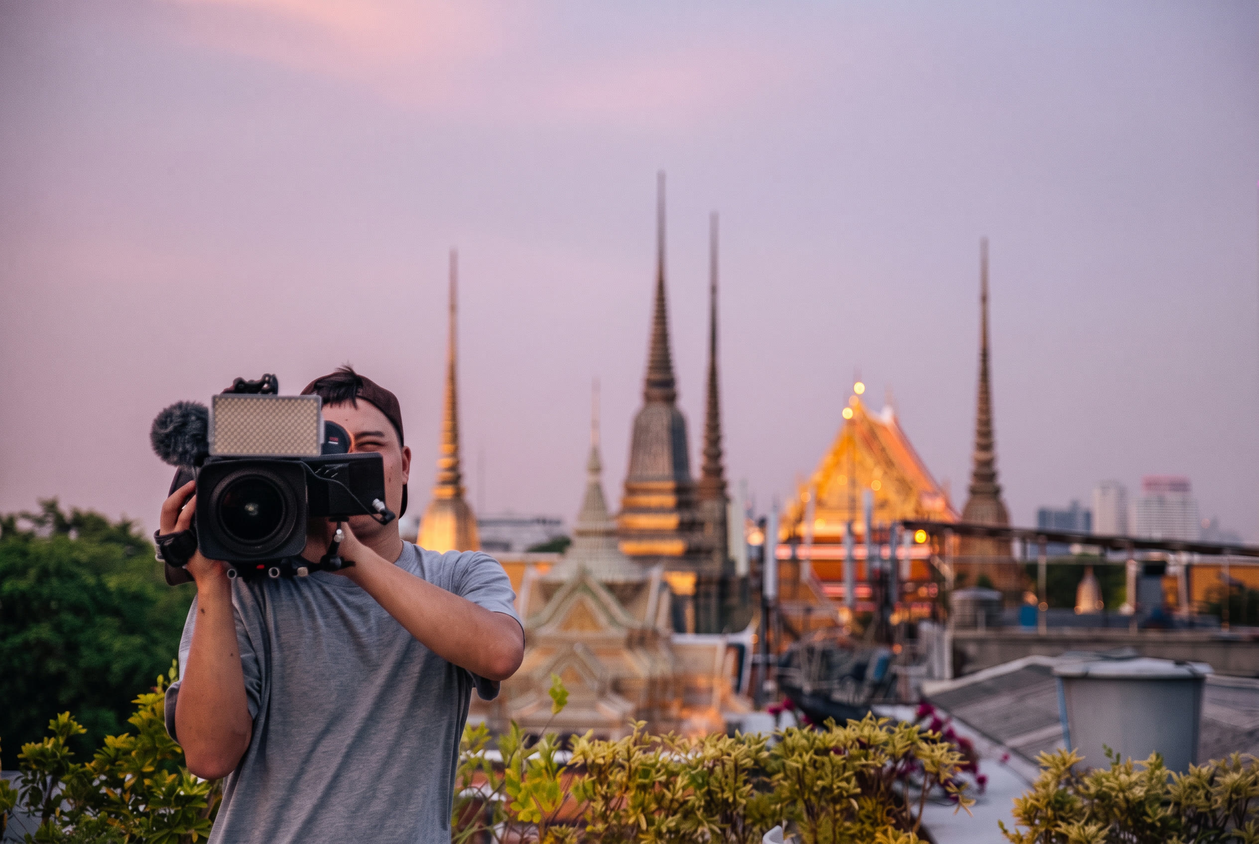 Cameraman filming at dusk with illuminated Bangkok temple spires in the background